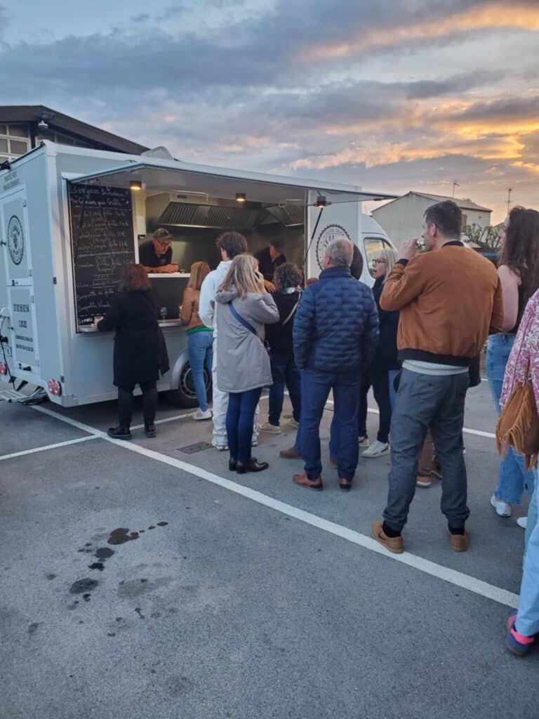 Food Truck au Crépuscule : File d'Attente et Menu Clientèle faisant la queue devant un food truck gris au crépuscule ; menu lisible : "Bonjour bienvenue au camion qui grille".