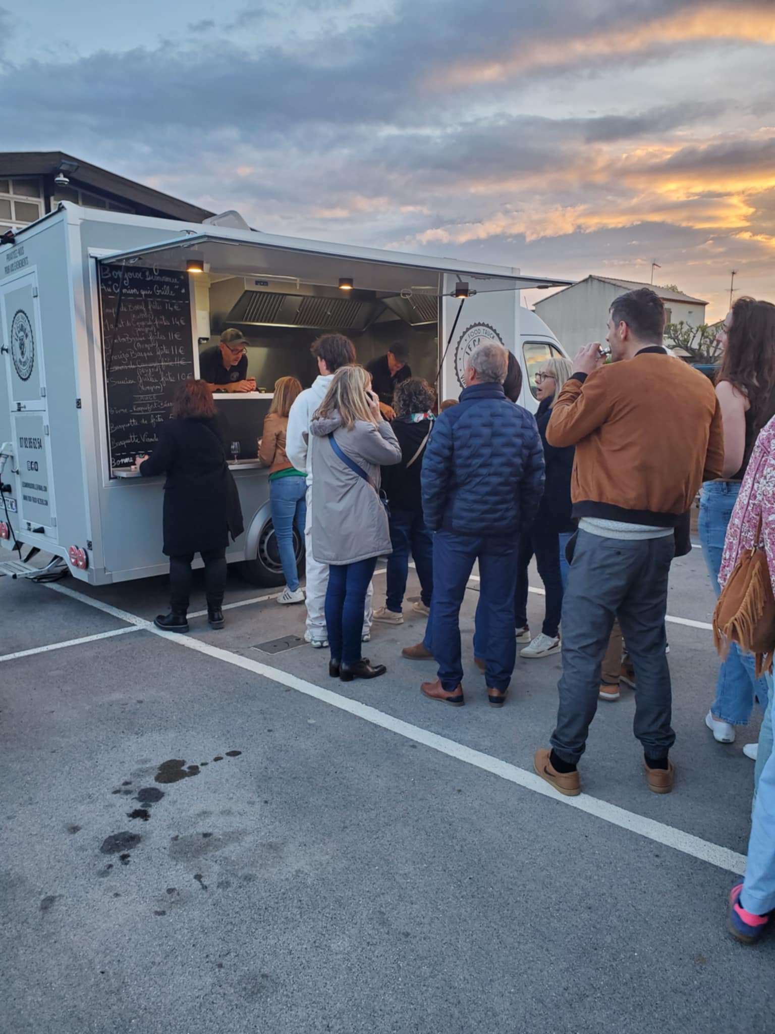 Clientèle faisant la queue devant un food truck gris au crépuscule ; menu lisible : "Bonjour bienvenue au camion qui grille".