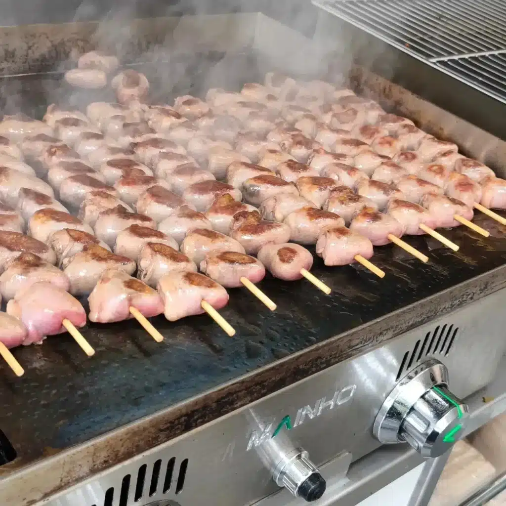 Brochettes de Saucisses Grillées à la Vapeur sur Plaque MAINHО Brochettes de saucisses ou de viande rosée grillant sur une plancha professionnelle MANINHO, dégageant de la vapeur.