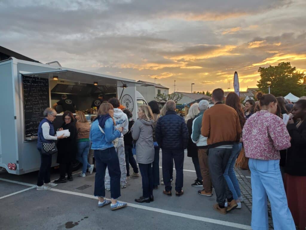 Food Truck « LE CAMION » au Coucher du Soleil Foule attendant des plats devant un food truck « LE CAMION » au coucher du soleil. Le menu indique des prix comme 14€ et 11€.