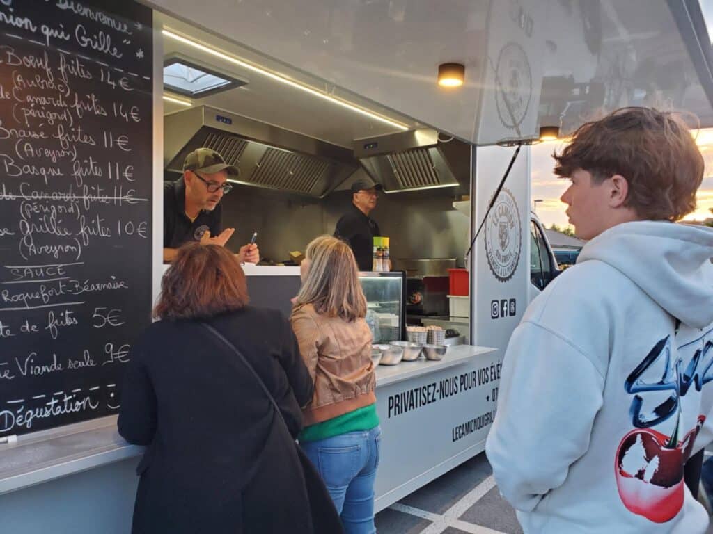 Food Truck Le Camion Qui Grille : Commandes et Menu Clients commandant dans un food truck 'Le Camion Qui Grille' avec un menu affiché : Boeuf frites 14€, Canard frites 14€, Sauces Roquefort-Béarnaise 5€.