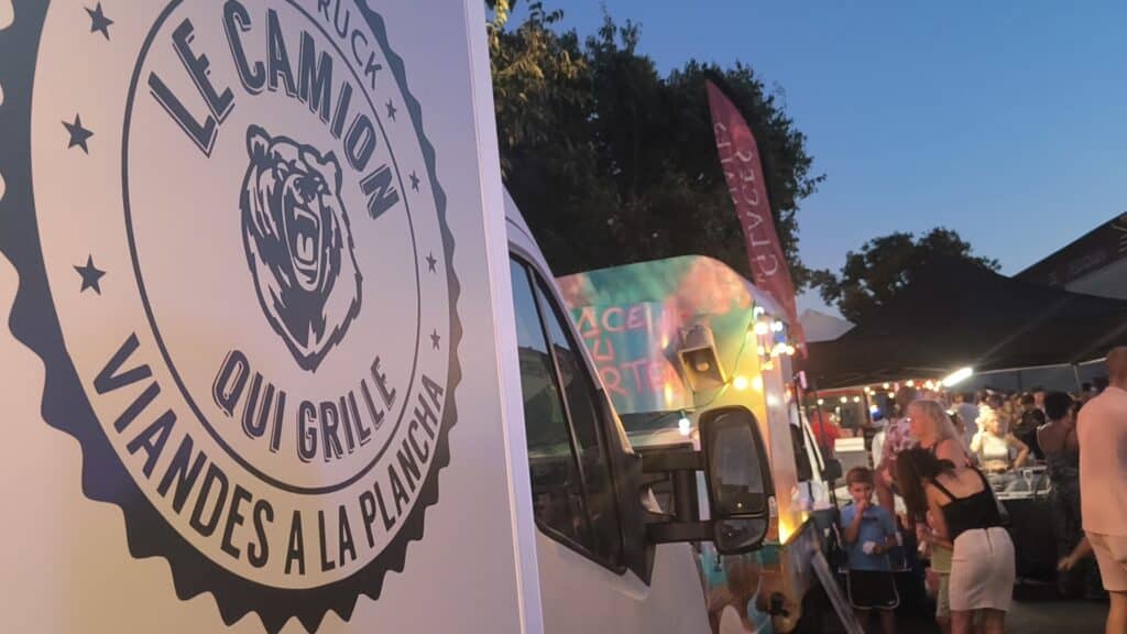 Food Truck Le Camion Qui Grille Viandes Panneau du food truck "LE CAMION QUI GRILLE VIANDES A LA PLANCHA" avec logo d'ours devant une foule en soirée.