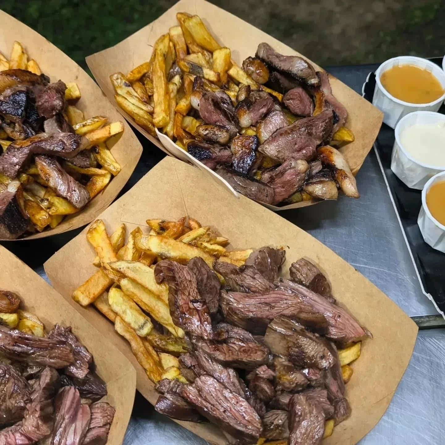 Quatre barquettes de frites dorées garnies de tranches de steak grillé, accompagnées de coupelles de sauces jaune et blanche.